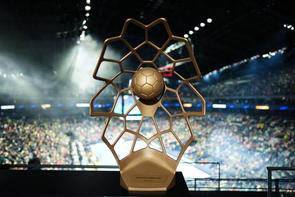 A golden trophy featuring a soccer ball encased in a hexagonal lattice design is displayed in a stadium filled with spectators.