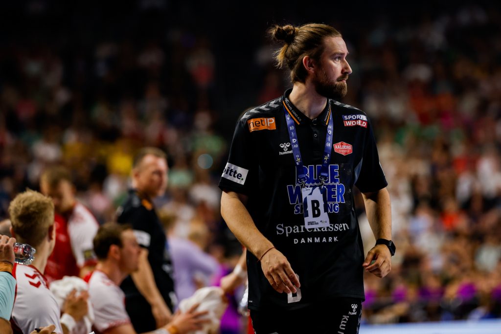 A person with a beard and hair tied in a bun stands on a sports court wearing a black jersey with sponsor logos and a lanyard, surrounded by a blurred crowd.