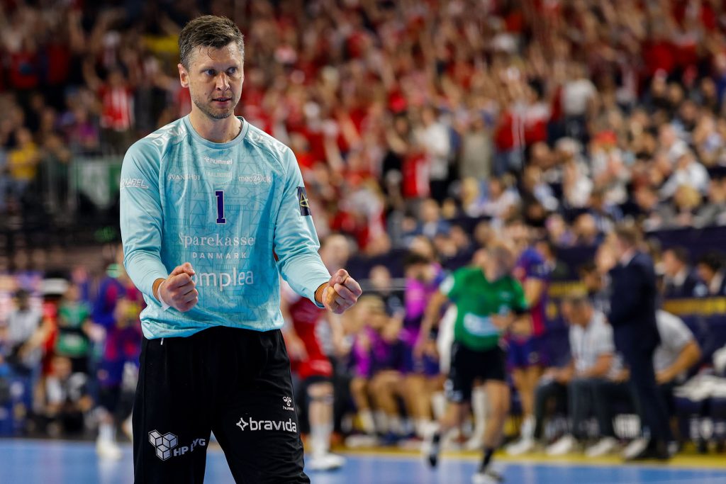 A handball player in a light blue jersey with the number 1 stands on the court, facing forward, with a crowd of spectators in the background.