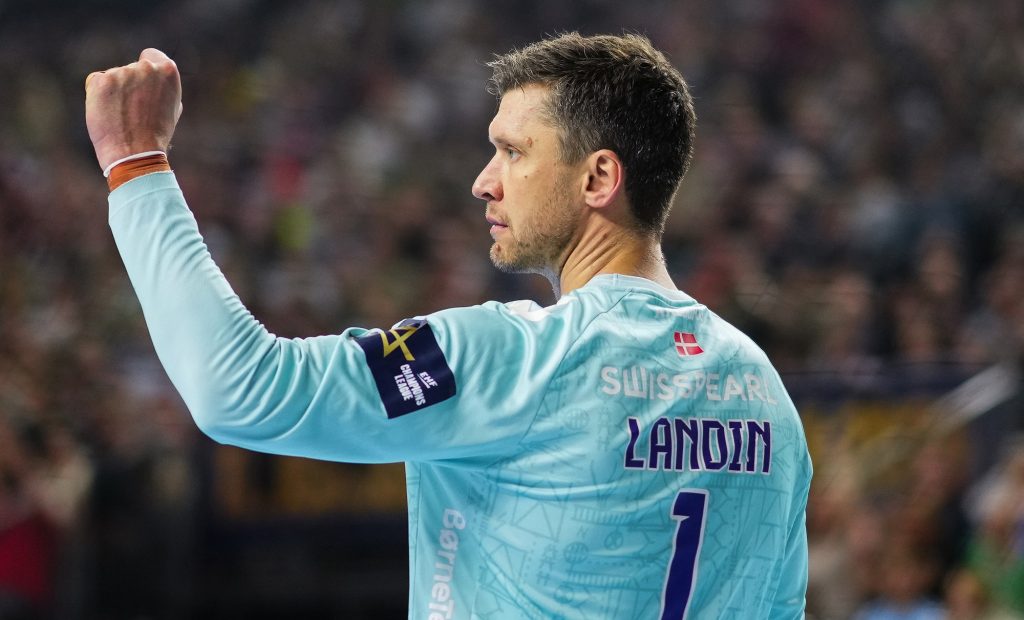 A goalkeeper in a light blue jersey with the number 1 raises his fist in celebration during a handball match.